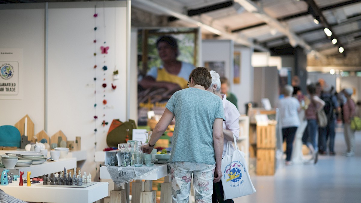 Zwei Besucherinnen schauen sich Produkte an einem Messestand an. Mehr Besucher*innen und Stände sind im Hintergrund.