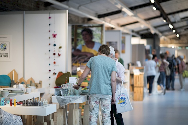 Zwei Besucherinnen schauen sich Produkte an einem Messestand an. Mehr Besucher*innen und Stände sind im Hintergrund.
